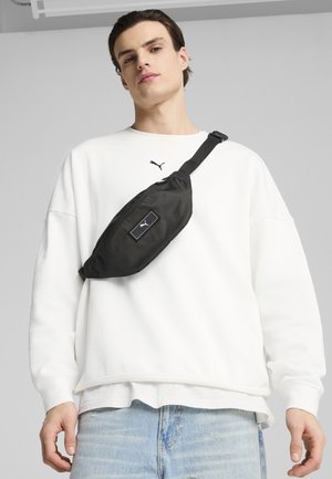 Black waist bag with textured material, featuring a prominent logo patch. Worn over a white sweatshirt and paired with light blue jeans.