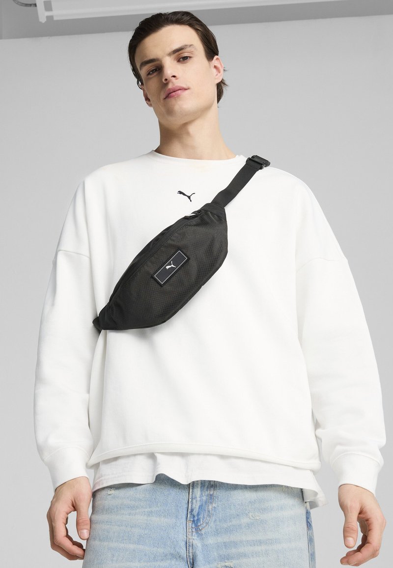 Svart midjeväska med texturerat material, prydd med en framträdande logotyp-patch. Bärs över en vit sweatshirt och matchas med ljusblå jeans.