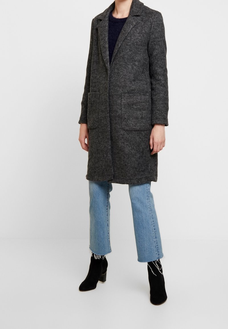 Cappotto di lana grigio con colletto, due tasche frontali e maniche lunghe, abbinato a jeans blu cropped e stivali neri alla caviglia. Texture liscia.