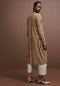 Kardigan berwarna coklat panjang dengan pola tekstur, dipadukan dengan celana pendek beiges terang dan sepatu mules bertumit abu-abu terang. Desain minimalis.