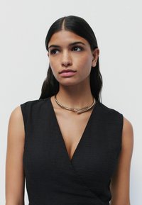 Gul chokerhalsband med en slät metallfinish, som har en minimalistisk design med kurviga detaljer, bärs över en svart topp.
