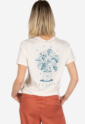 Person with shoulder-length blonde hair wearing a white T-shirt with a blue plant illustration and "STAY DIFFERENT" text on the back.