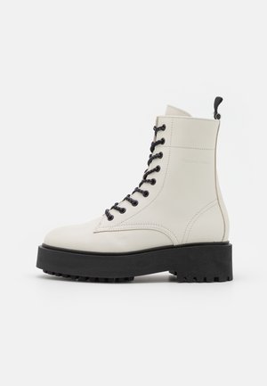 Botín de tobillo en cuero blanco con cordones negros, suela de plataforma de goma y patrón de huella texturizado. Lengüeta de acento en el talón.