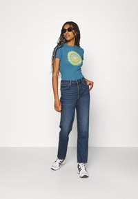 T-shirt bleu à manches courtes avec un graphique de soleil jaune et vert, associé à un jean bleu foncé taille haute et des baskets blanches.