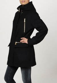 Zwarte jas met capuchon, voorzien van gouden ritssluitingaccenten en twee ritszakken. Gemaakt van een gestructureerd materiaal, reikt tot onder de heupen.