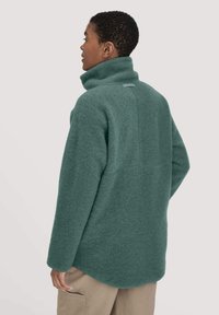 Fleece pullover in soft teal with a high collar, relaxed fit, and a back seam detail. Features a subtle tag near the neck.