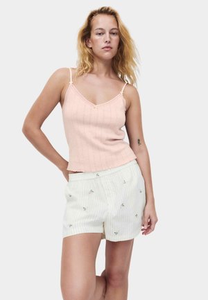Kvinde med blondt hår iført en lyserød ærmeløs top og hvide, stribede shorts med små blomsterprint mod en ensfarvet baggrund.