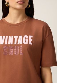 Kobieta w brązowym T-shircie z napisem "VINTAGE SOUL", złotych kolczykach w kształcie kół, z zamkniętymi ustami i prostymi, ciemnymi włosami.