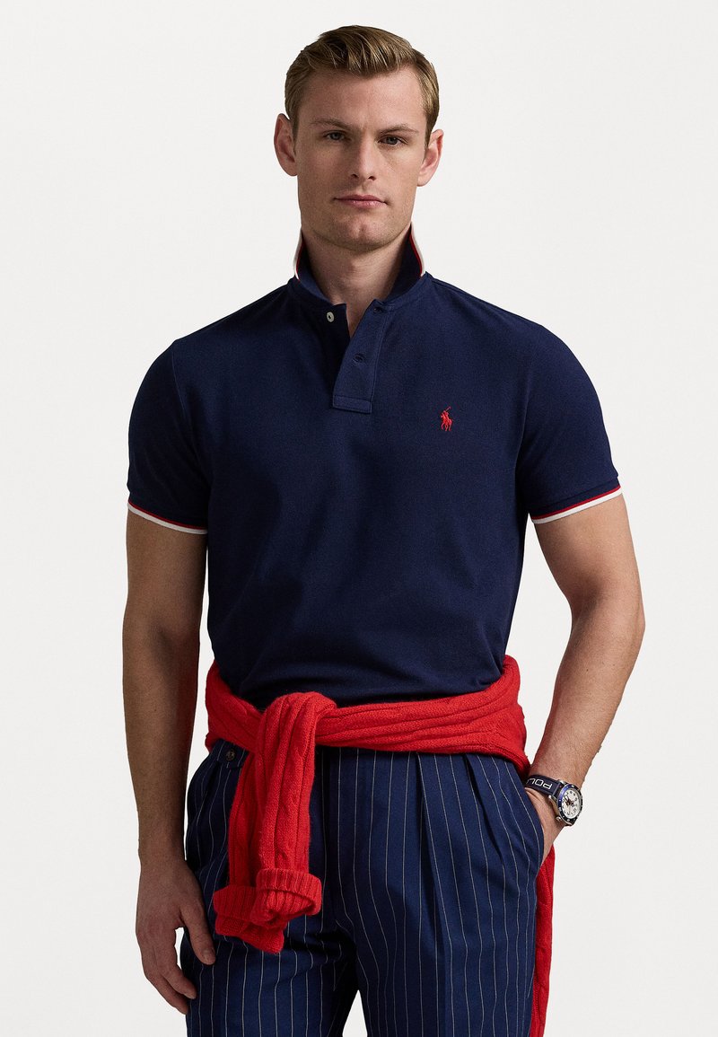 Polo de marinha com um fecho de dois botões e logótipo vermelho, apresentando detalhes em riscas vermelhas e brancas na gola e nas mangas. Usado com um suéter vermelho.