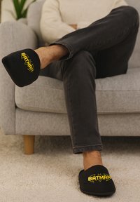 Pantofole nere con una texture morbida, caratterizzate da un logo giallo "BATMAN" e dal simbolo del pipistrello sulla parte anteriore. Indossate ai piedi di una persona.