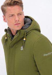Veste à capuche vert olive avec un extérieur lisse, doublure en polaire grise, fermeture éclair et cordons contrastés. Présente un logo sur la poitrine.