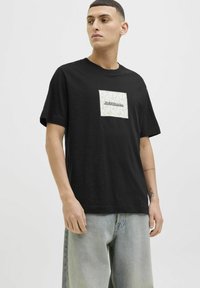 Juodos medvilnės marškinėliai su balta ir auksine kvadratinėmis aplikacijomis, kuriose pavaizduotas "Jack & Jones" logotipas, trumpomis rankovėmis ir klasikinio apvalaus kaklo dizainu.