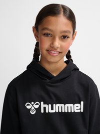 Jeune fille aux cheveux tressés portant un sweat à capuche noir avec un logo blanc « hummel » et un symbole d'abeille, souriant doucement sur un fond uni.