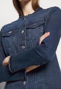 Chaqueta de mezclilla azul oscuro con cierres de botón, bolsillos en el pecho y cuello y puños deshilachados. Presenta un diseño ajustado y costuras visibles.
