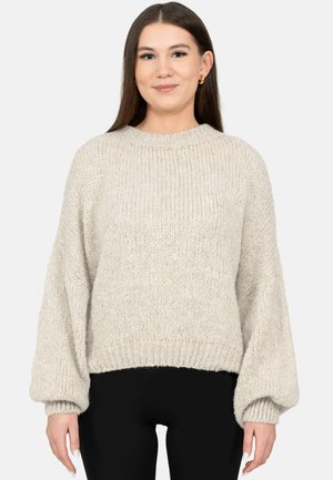 Young woman with long brown hair wearing a light gray chunky knit sweater and black pants, smiling slightly, against a white background.