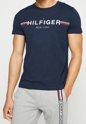 Mężczyzna ubrany w granatowy T-shirt z napisem "Hilfiger New York" oraz szare spodnie dresowe z paskiem z logo Tommy Hilfiger na lewej nodze.