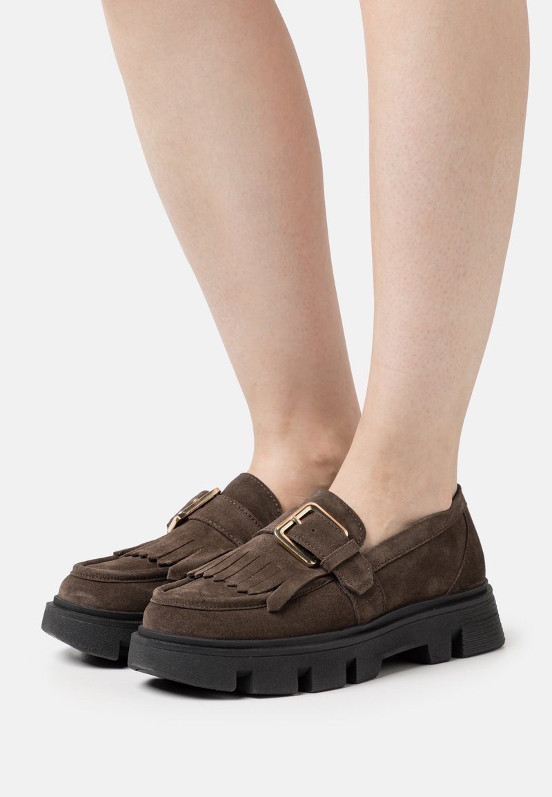 Mocassins en daim marron avec franges et détail de boucle dorée, portés sur des jambes à la peau claire, avec des semelles épaisses en caoutchouc noir texturé.