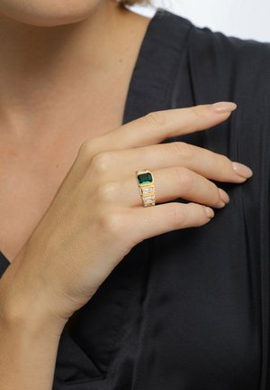 Guldring med en stor grøn rektangulær sten og tre mindre klare sten, sat på en tekstureret ring, båret på en hånd.