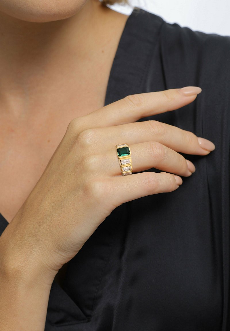 Goldring mit einem großen grünen rechteckigen Stein und drei kleineren klaren Steinen, auf einem strukturierten Band, getragen an einer Hand.