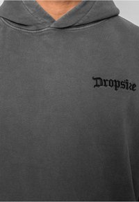 Grå hættetrøje med en blød tekstur, der har en frontlomme og et sort broderet "Dropsie" logo på venstre brystområde.
