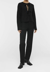 Pull noir en maille avec un col en forme de goutte et deux nœuds à l'avant, associé à un pantalon noir droit et des bottines.