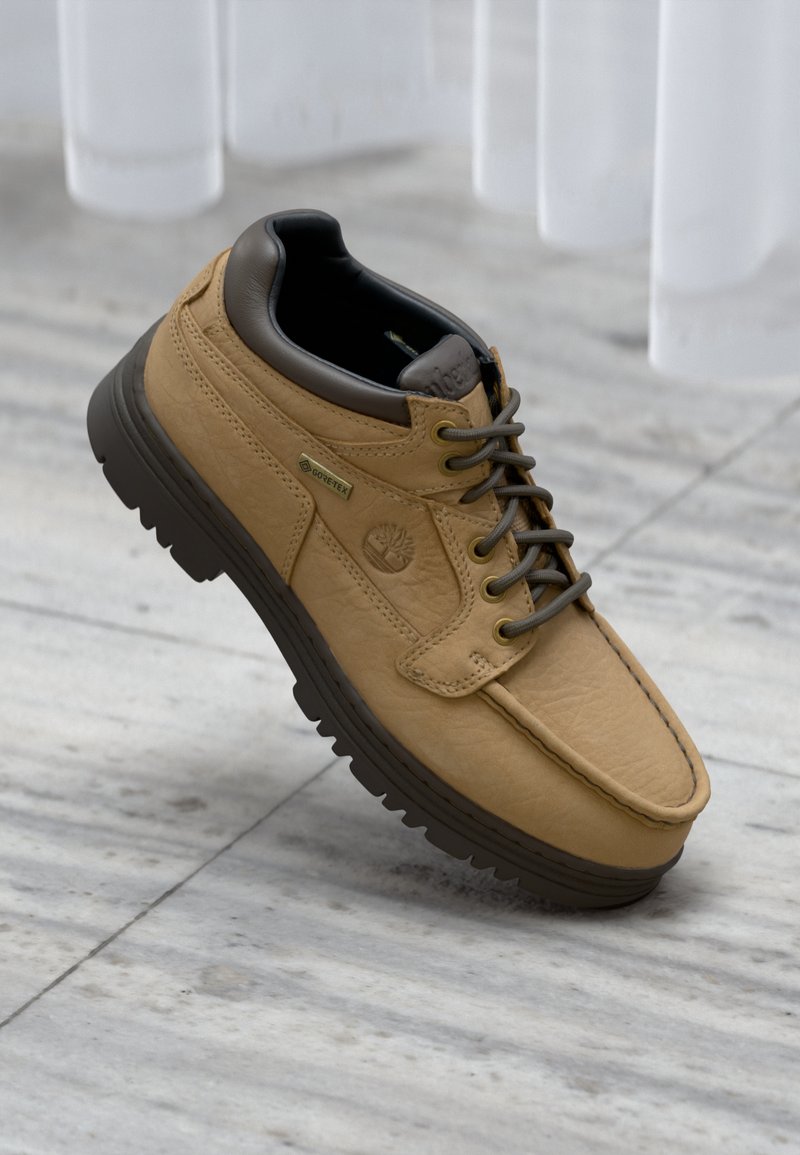 Chaussure en cuir marron clair à lacets avec semelle et col foncés, étiquette Gore-Tex et logo Timberland sur fond de sol en marbre.