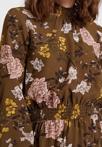 Kleid mit Blumenmuster aus braunem Stoff, verziert mit rosa und gelben Blumen. Geraffter Ausschnitt und Taillenbereich mit langen Ärmeln. Strukturierte Oberfläche.