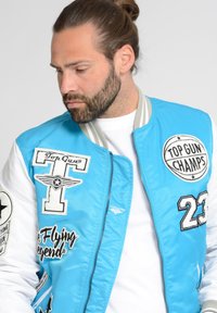 Blauwe bomberjack met witte mouwen, voorzien van geborduurde patches, waaronder "Top Gun" en "23," met ritssluiting en een relaxte pasvorm.