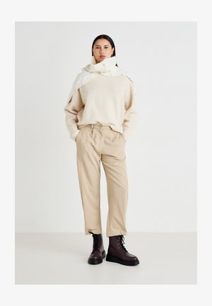 Maglione di lana beige con un colletto alto, sovrapposto a un leggero wrap crema; pantaloni beige larghi; stivali marrone scuro con lacci e suola spessa.