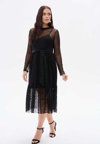 Czarna sukienka z długimi transparentnymi rękawami, dopasowanym korpusem i warstwową koronkową spódnicą w wzór lamparta. W komplecie pasek w talii i sandały na paski.