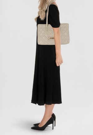 Borsa a tracolla beige imbottita con due manici e dettaglio del logo color oro, abbinata a un vestito nero texturizzato e tacchi alti neri.