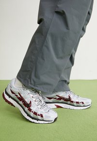 Vita och grå sportskor med röda accenter, nätpaneler och en texturerad sula, använda med grå byxor på en grön yta.