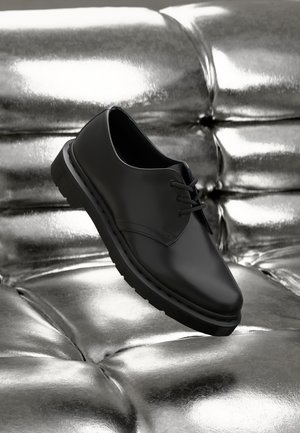 Chaussure noire en cuir à lacets avec un bout arrondi, une semelle en caoutchouc et un design minimaliste, présentée sur un fond métallique brillant.