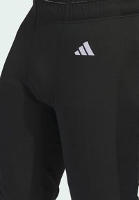Black athletic pants made of stretchy material, featuring an elastic waistband and a white logo on the left thigh. Smooth texture.