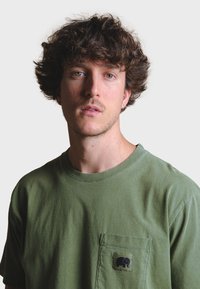 Green cotton t-shirt featuring a round neckline and a left chest pocket with a black elephant logo. Smooth texture, casual design.