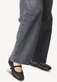 Schwarze Lederschuhe mit abgerundeter Spitze, ausgestattet mit einem Riemen über dem Spann. Getragen mit weit geschnittenen, dunkelgrauen Jeans, die die flache Sohle der Schuhe zeigen.