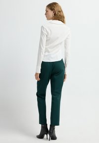 Witte blouse met lange mouwen en een kraag, gecombineerd met groene broek en zwarte hoge enkellaarsjes. Eenvoudig ontwerp, gladde textuur van de stof.