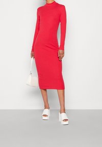 Personne portant une robe midi rouge ajustée à manches longues, tenant un petit sac à main blanc et des escarpins blancs à plateforme ouverts.