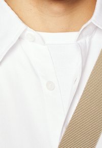 Vit skjorta med krage och knäppning fram, lager över ett vitt ribbat plagg, åtföljd av ett strukturerat beige bälte.