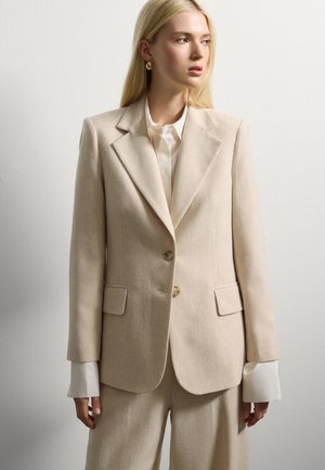 Donna bionda che indossa un blazer beige su misura e pantaloni abbinati, con i polsini di una camicia bianca visibili, in piedi davanti a un muro bianco semplice.