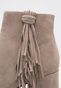 Botín de tobillo de ante beige con un acabado texturizado, que presenta un detalle de borlas anudadas hechas de flecos finos en la parte trasera.