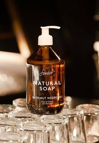 Bottiglia di vetro marrone di sapone naturale con pompetta bianca, etichettata "Sapone Naturale" e "Wermut Negroni", poggiata su contenitori di vetro trasparente.