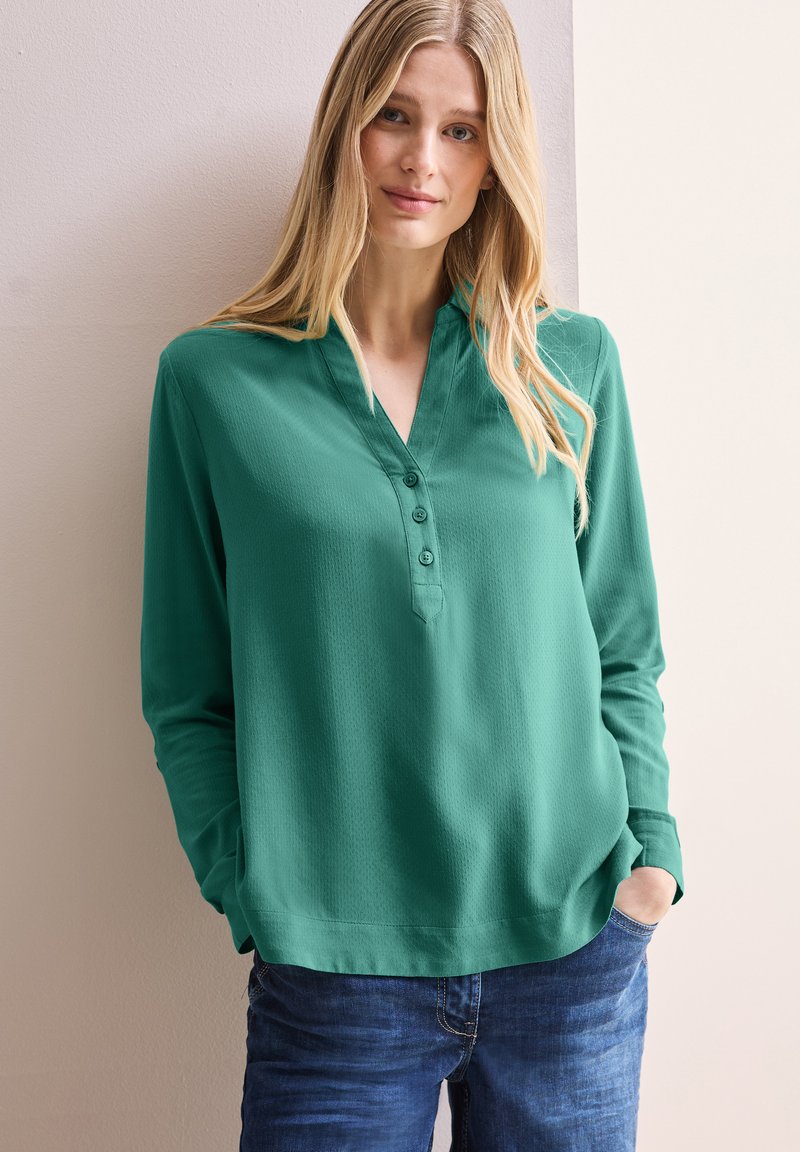 Blusa verde a manica lunga con scollatura a V, tre bottoni e tessuto testurizzato. Abbinata a jeans blu, presenta una vestibilità rilassata.