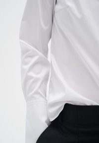 Personne portant une chemise blanche à manches longues avec de larges poignets, associée à un pantalon noir, montrée du milieu du torse au poignet.