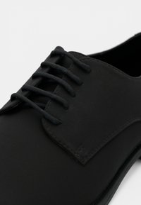 Chaussure Oxford en cuir noir avec laçage fermé et lacets ronds noirs sur fond blanc.
