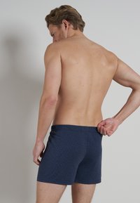 Aansluitende zwemshorts in donker marineblauw met een gestructureerde ruitpatroon. Elastische tailleband, lengte tot halverwege de dij en geen zichtbare sluitingen of accenten.