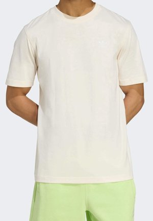 Mężczyzna ubrany w prosty jasnobeżowy T-shirt z krótkim rękawem, z małym białym logo Adidas na lewej piersi, w połączeniu z jasnzielonymi szortami.