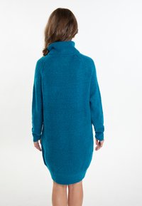 Rochie tricotată supradimensionată de culoare teal, cu guler înalt, mâneci lungi și finisaj texturat; ușor conică la margine.