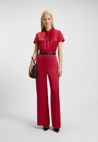 Incostume rosso con una parte superiore a colletto e maniche corte, e pantaloni larghi, cinte in vita. Presenta una texture liscia e una borsa nera.