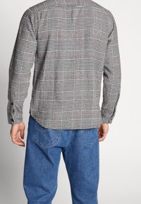 Man som bär en grå rutig långärmad skjorta och blå jeans, sedd bakifrån mot en enfärgad bakgrund.